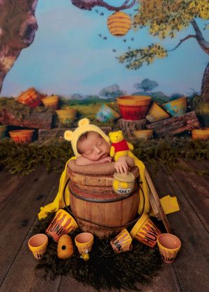 Sleeping newborn in yellow Winnie the Pooh bonnet posed in a wooden barrel with honey pot props.