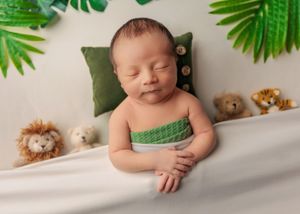 Peaceful sleeping newborn swaddled in green on a white bed surrounded by jungle animal plush toys.