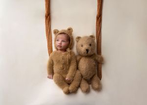 Sleeping newborn dressed in a brown bear outfit beside a matching teddy bear on a swing.