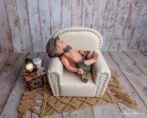 Newborn baby posed in miniature armchair with vintage props on a rustic wooden floor backdrop.