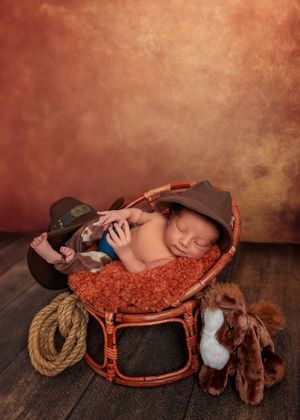 Sleeping newborn in cowboy hat nestled in a wicker basket with rope and toy horse on wood floor.