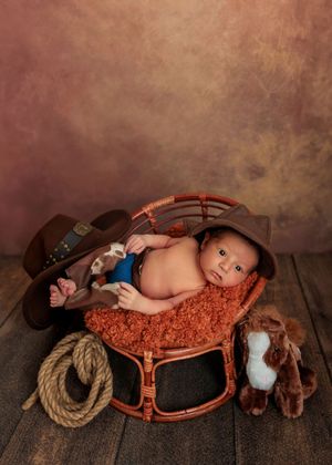 Alert newborn baby in cowboy hat posed in a wicker basket with rope and toy horse props.