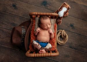 Sleeping newborn baby in blue diaper posed in a rustic wooden rack with cowboy hat and rope props.