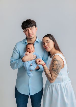 Young parents in light blue outfits proudly holding their newborn baby in a studio portrait photo.