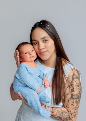 Young tattooed mother in blue dress gently holding her newborn baby in a studio portrait photo.