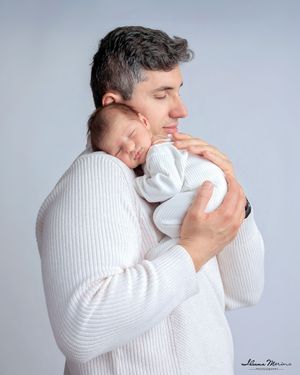 Father in white sweater tenderly holding sleeping newborn baby against his chest in studio portrait.
