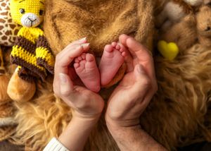 Parent hands cradling tiny newborn baby feet surrounded by stuffed animal toys on a fur blanket.