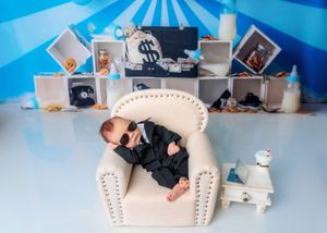 Newborn baby in black suit and sunglasses posed in a white armchair with money bag themed backdrop.