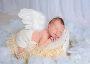 Sleeping newborn baby posed with white angel wings on a fluffy cream blanket in a small bucket.