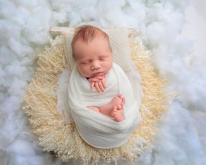 Sleeping newborn wrapped in white swaddle with tiny feet peeking out, posed in a fluffy yellow basket.