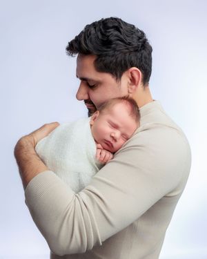 Father in beige turtleneck tenderly cradling his sleeping swaddled newborn baby against his chest.