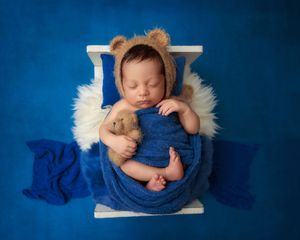 Newborn baby in bear ear bonnet sleeping with teddy bear on white bed with blue blanket backdrop.