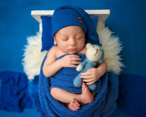 Newborn baby sleeping in blue wrap and hat holding a knit teddy bear on a small white bed.