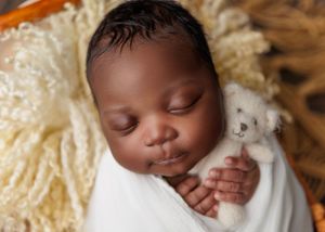 Sleeping newborn swaddled in white wrap holding a small white teddy bear in a cream-lined basket.