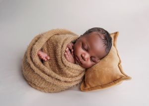 Sleeping newborn wrapped in tan knit swaddle resting on a matching brown suede pillow.