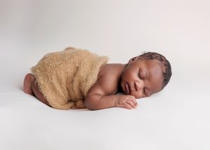 Peaceful sleeping newborn wrapped in a tan knit blanket lying on a soft white surface.