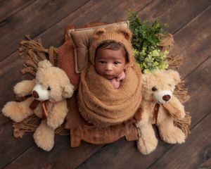 Swaddled newborn in bear hat on tiny bed flanked by two teddy bears.