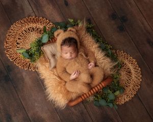 Sleeping newborn in brown teddy bear outfit resting in wicker basket with greenery on wood floor.