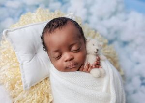 Sleeping newborn baby swaddled in white wrap holding a tiny teddy bear on a yellow fluffy blanket.