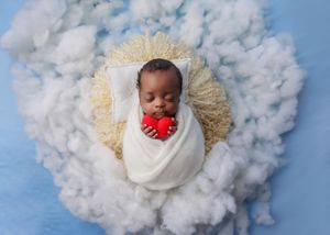 Newborn baby wrapped in white swaddle holding a red heart while sleeping in a cloud-themed photo setup.