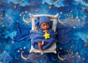 Newborn in blue wizard hat holding yellow star sleeping on tiny bed with celestial backdrop.