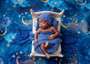 Sleeping newborn wrapped in blue blanket with star hat on miniature bed against starry night backdrop.