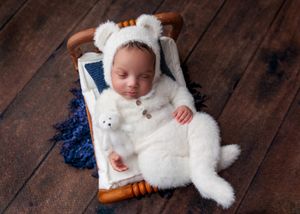 Sleeping newborn in a white bear outfit holding a stuffed animal on a wooden bed.