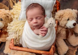 Swaddled newborn baby sleeping in wicker basket holding small teddy bear surrounded by stuffed animals.