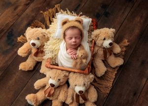 Sleeping newborn wrapped in white swaddle in small wooden bed surrounded by teddy bears on wood floor.