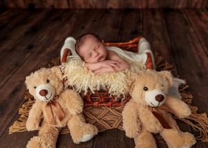 Peacefully sleeping newborn baby in basket with fluffy lining flanked by two teddy bears on wooden floor.