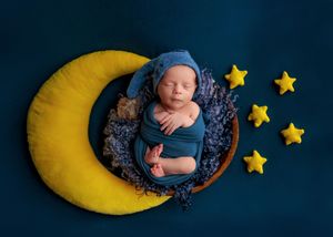 Swaddled newborn baby in blue wrap sleeping in wooden bowl on yellow moon prop with yellow stars.