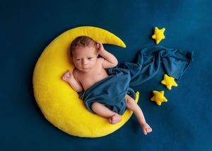 Newborn baby lying on yellow crescent moon pillow with teal blanket and yellow star props on blue background.