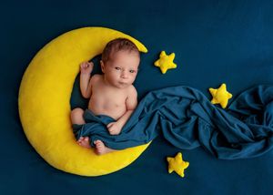 Newborn baby resting on yellow crescent moon prop with teal blanket and star decorations on blue background.