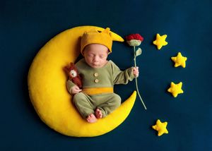 Sleeping newborn in green outfit holding flower on yellow moon prop with teddy bear and stars on blue background.