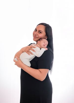 Beautiful dark-haired mother in black dress holding sleeping newborn baby in white studio photo.