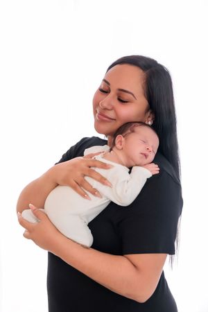 Smiling mother in black dress tenderly holding her sleeping newborn baby on white background.