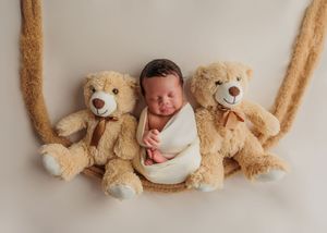 Adorable newborn baby wrapped in white swaddle posed between two teddy bears on a swing.