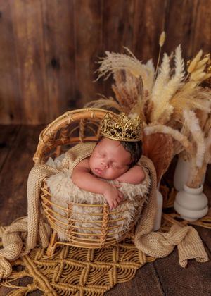 Sleeping newborn baby wearing a gold crown in a wicker basket with boho pampas grass decor.