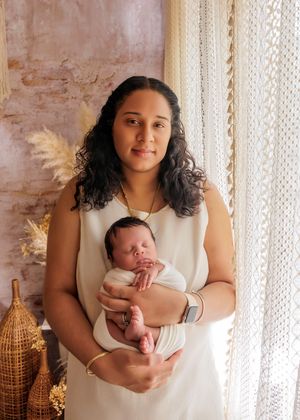 Young mother in white dress tenderly holding her sleeping newborn baby in a boho studio setting.