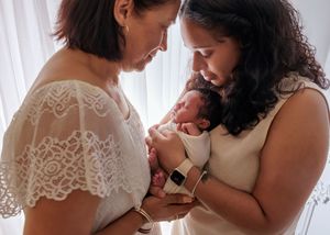 Two women lovingly gazing at a newborn baby they are holding together near a bright window.