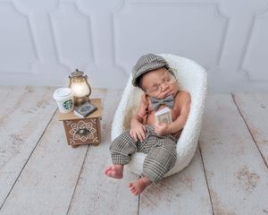 Newborn baby sleeping in tiny chair wearing plaid newsboy cap and suspenders beside miniature Starbucks cup.
