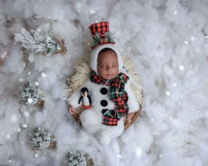 Newborn dressed as a snowman in a basket surrounded by snow and pine trees.