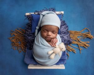 Sleeping newborn swaddled in blue with a tiny teddy bear on a blue bed.