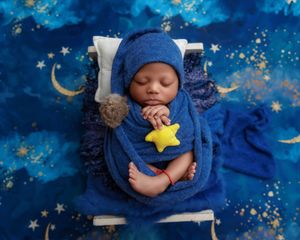 Newborn baby wrapped in blue swaddle holding a yellow star toy on a starry night-themed backdrop.