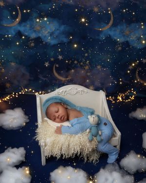 Sleeping newborn baby in blue outfit with teddy bear on white bed against a starry night sky backdrop.