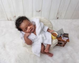 Smiling newborn in white robe relaxing with a mug on a fluffy white rug.