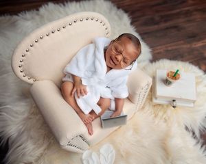 Smiling newborn baby in white robe and glasses relaxing on a miniature cream armchair with accessories.