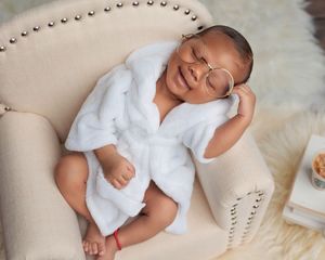 Smiling newborn baby in white fluffy robe wearing round glasses lounging on a small cream armchair.
