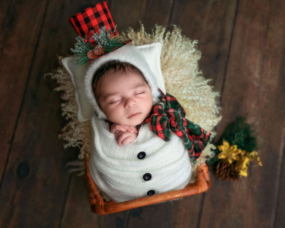 Sleeping newborn wrapped in white knit snowman swaddle with plaid top hat in rustic wicker basket.