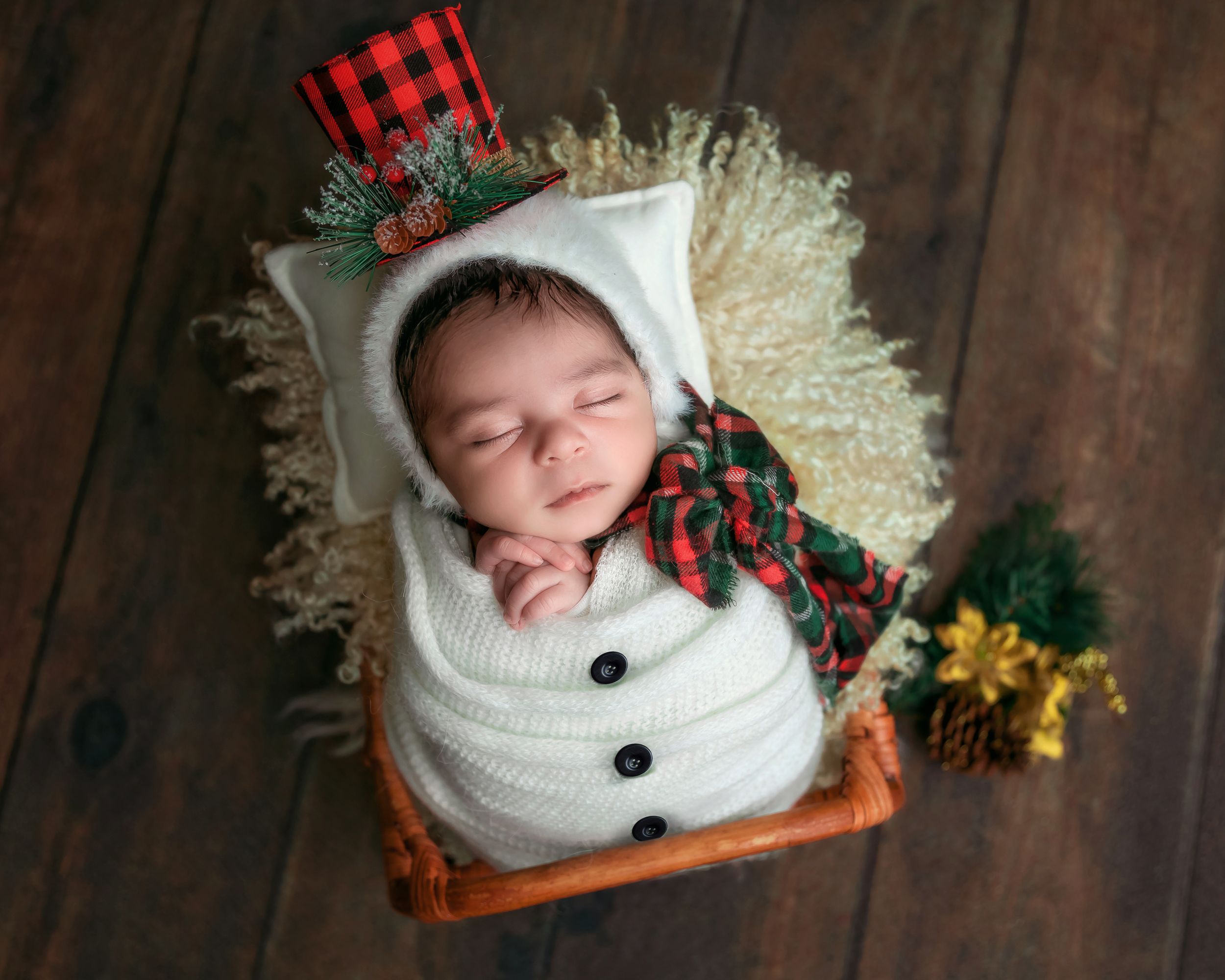 Sleeping newborn wrapped in white knit snowman swaddle with plaid top hat in rustic wicker basket.